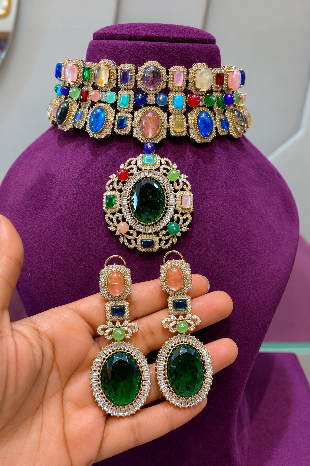 A multicolor gemstone jewelry set including a choker necklace, a matching pendant, and drop earrings, displayed on a purple velvet mannequin bust with a hand holding the earrings in front.