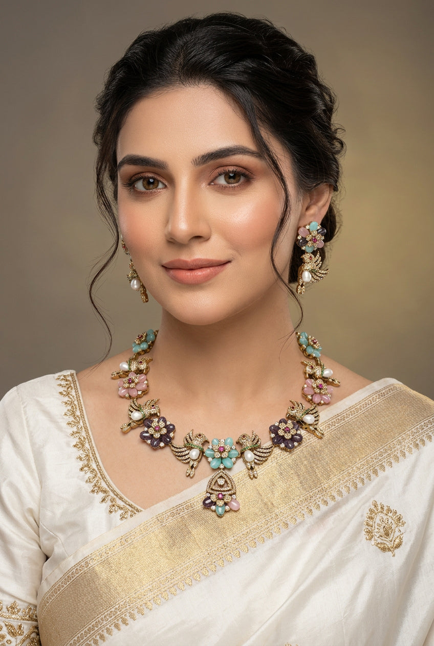 A gold-plated jewelry set featuring a necklace, earrings, and bracelet, each decorated with colorful enamel flowers and birds. The set includes turquoise, pink, purple, and white enamel details, displayed on a beige background with coffee beans and a wooden spoon prop.